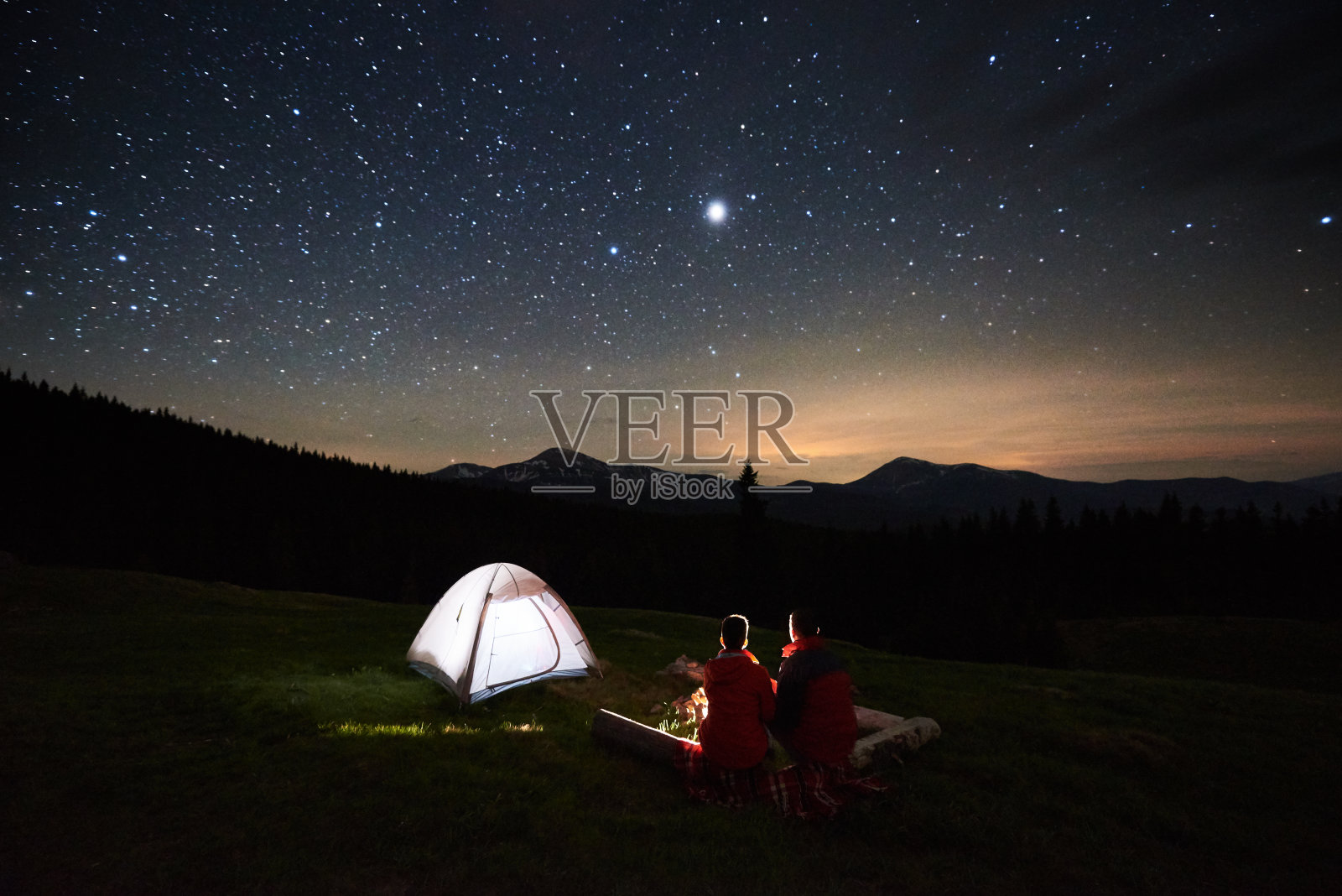 晚上露营。在夜空下，一对游客坐在篝火旁的剪影照亮了帐篷。天体摄影术照片摄影图片