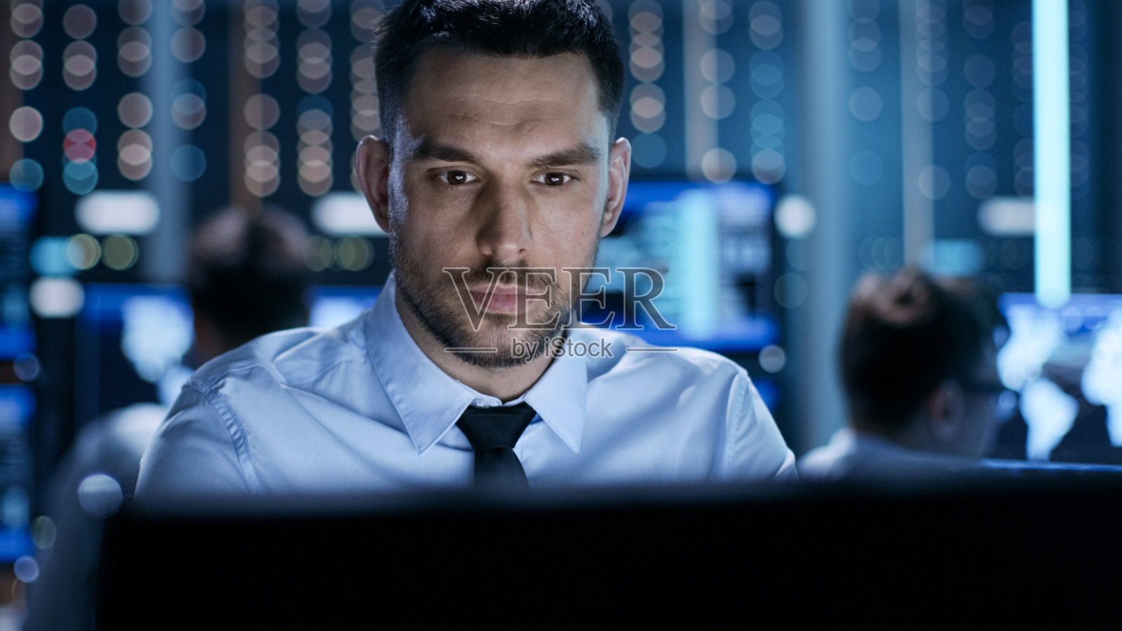 一个专业技术控制器坐在他的办公桌与多个显示在他之前的特写。在系统控制中心工作的同事。照片摄影图片
