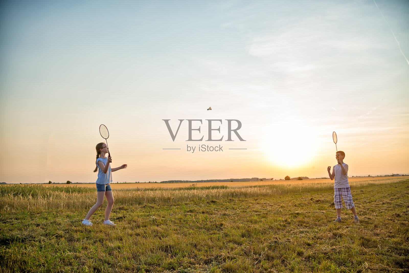 夏日日落时分，两个小孩在操场上打羽毛球照片摄影图片