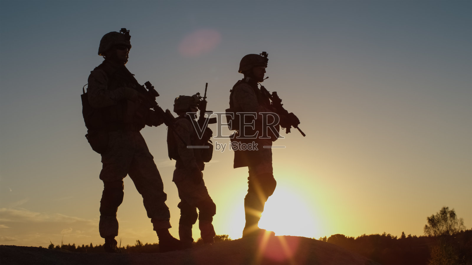 日落时分的沙漠环境中，三名全副武装的士兵站在山上。照片摄影图片
