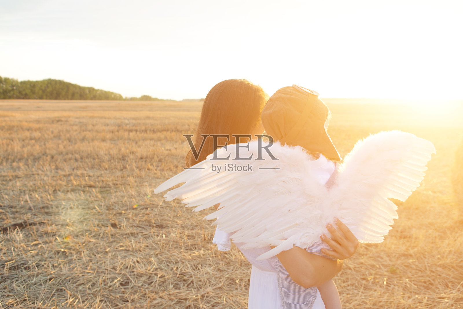 母亲和儿子玩得很开心。男孩戴着飞行员帽，带着天使翅膀坐在他妈妈的手上看日落。照片摄影图片