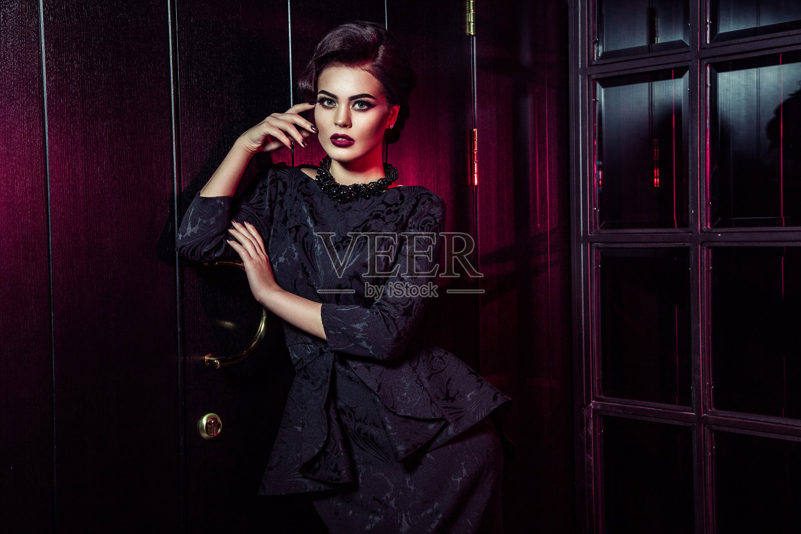 肖像美丽的时装模特在经典的黑色衣服，化妆和发型附近黑暗的门站着和摆姿势和从窗户的红色光在她身上。照片摄影图片