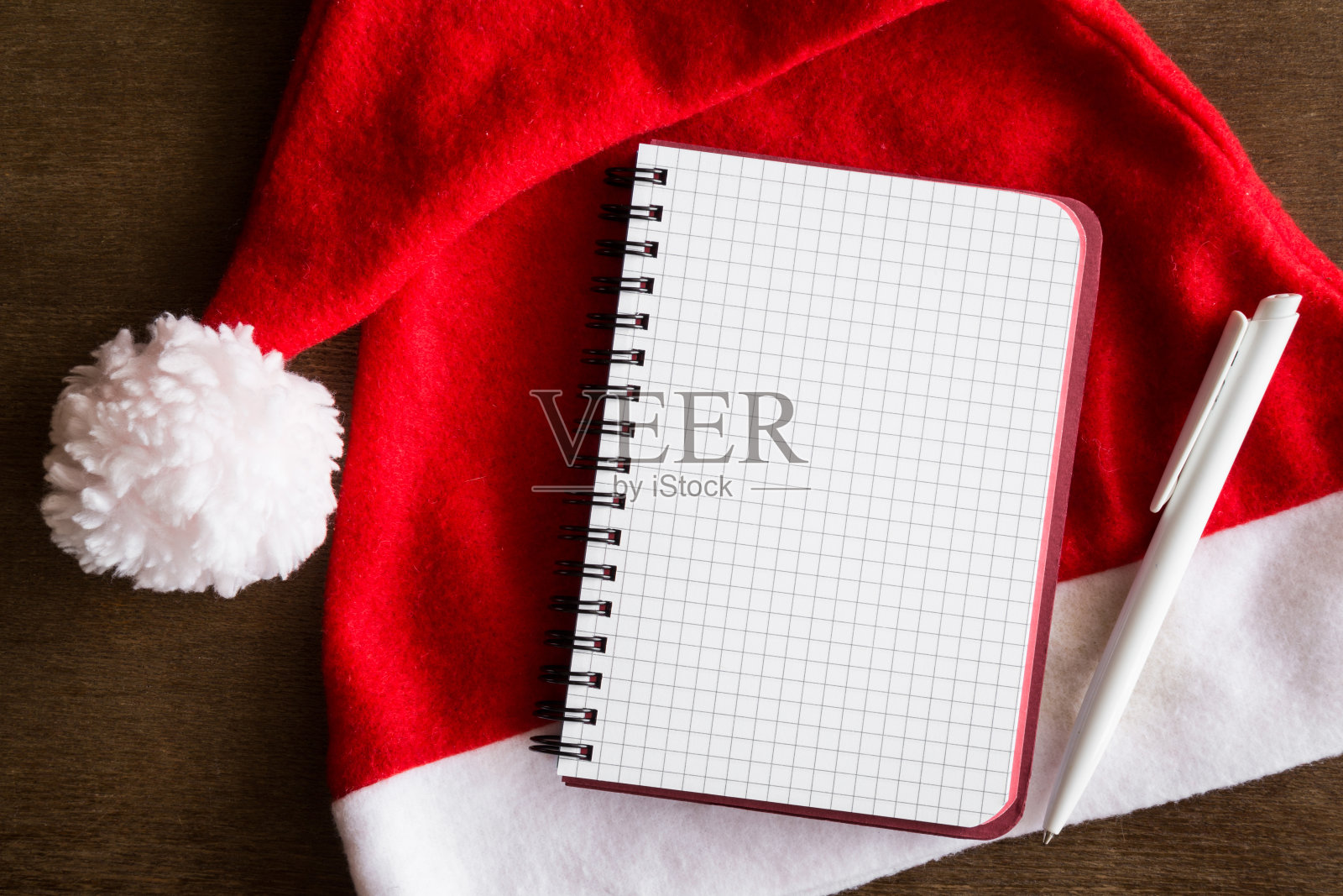 圣诞老人的红帽子，钢笔和笔记本放在木桌上。圣诞节的时间概念。为礼物清单或其他文字腾出地方。前视图。照片摄影图片