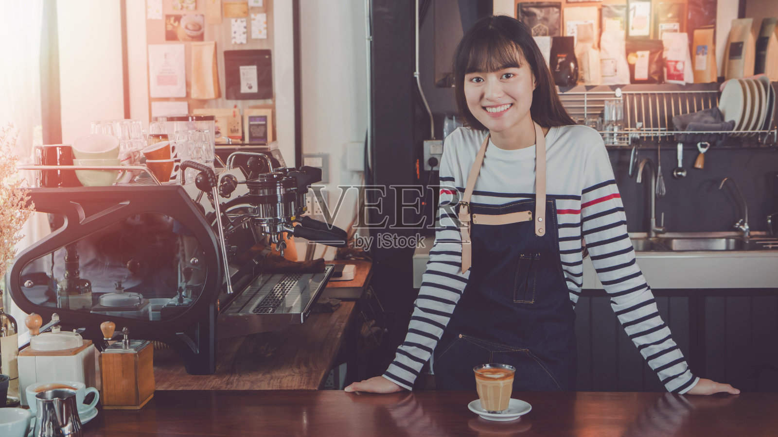 年轻美丽的亚洲女咖啡师，穿着蓝色围裙，拿着热咖啡杯，在咖啡店的吧台微笑着招待顾客。咖啡店和小型咖啡店的概念。的语气照片摄影图片