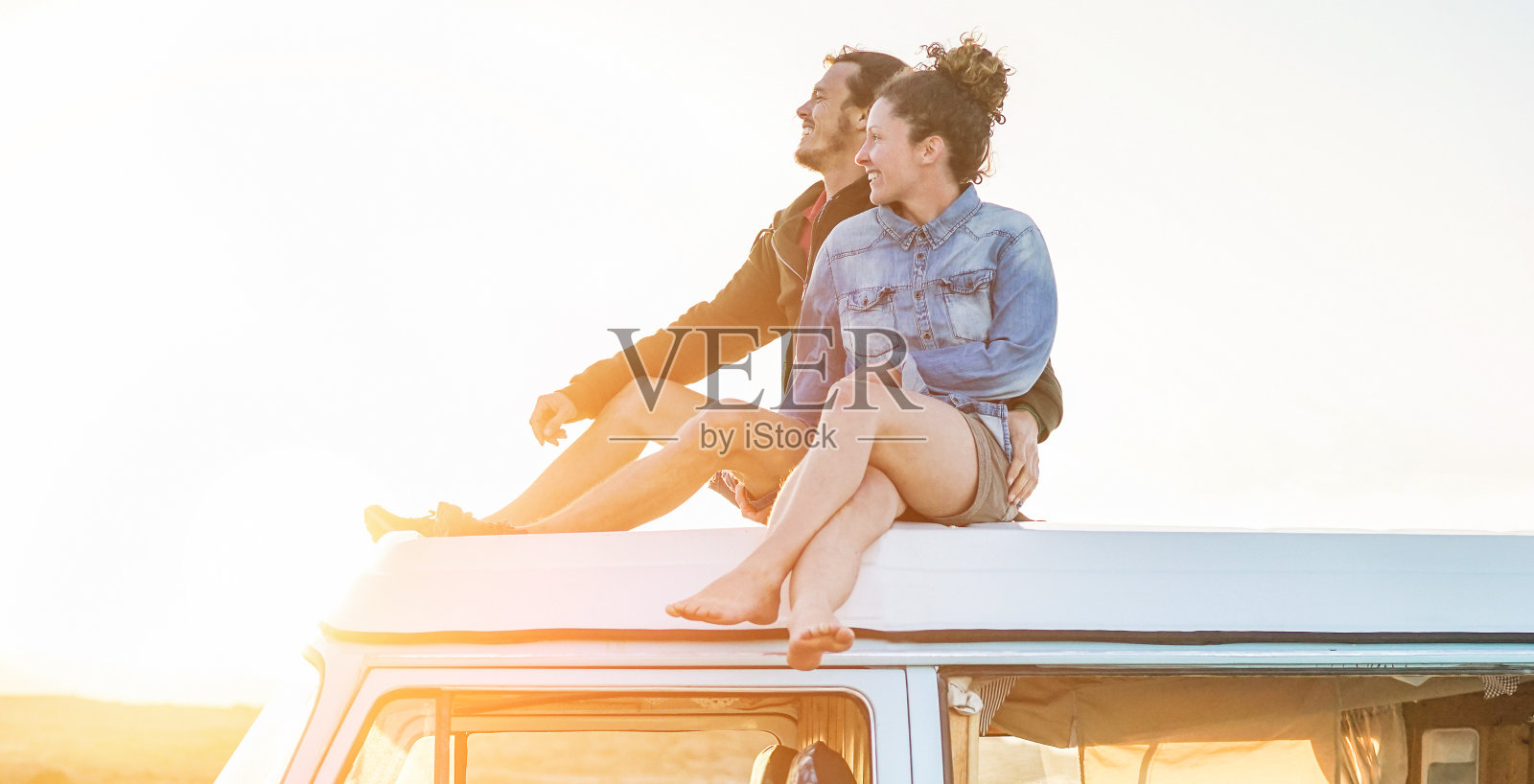 幸福的夫妇坐在小货车屋顶在日落-年轻人有乐趣的春假环游世界-旅行，爱和假日概念-关注脸照片摄影图片