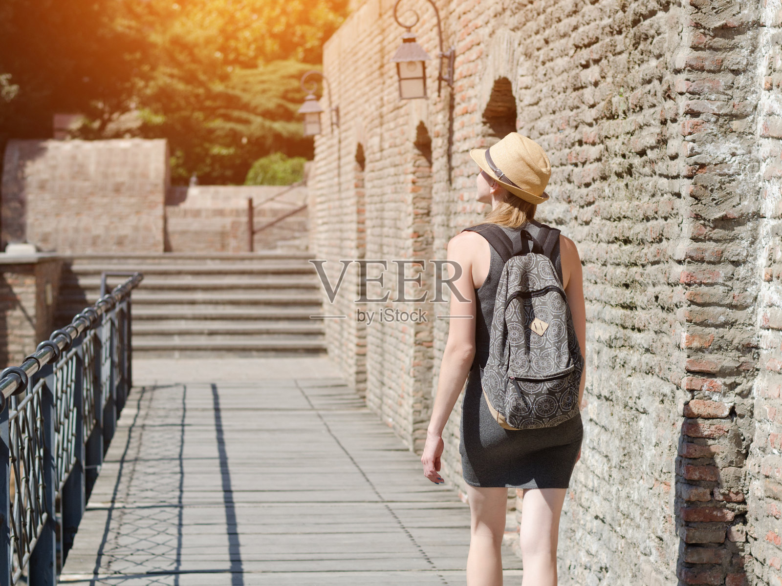 戴着帽子背着背包的女孩沿着城墙走着。阳光灿烂的日子照片摄影图片