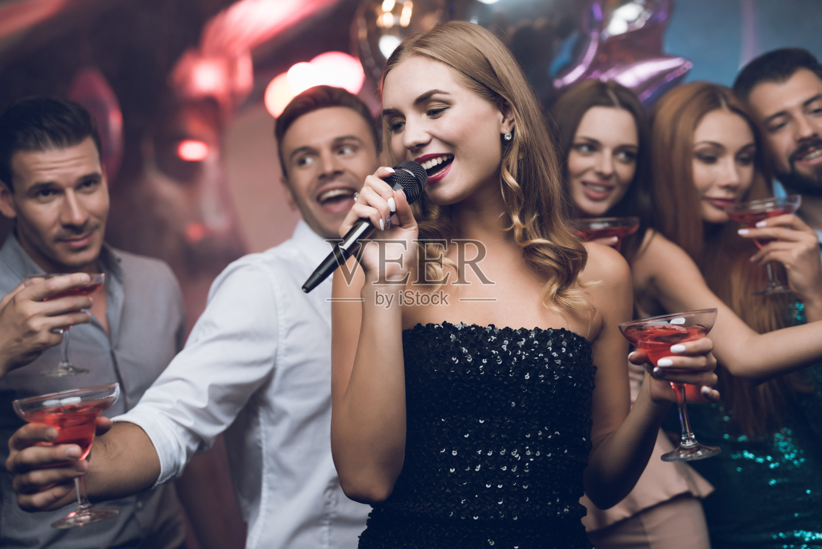 一个穿着黑裙子的女人正在和她的朋友在卡拉ok俱乐部唱歌。她的朋友们在后台玩得很开心。照片摄影图片