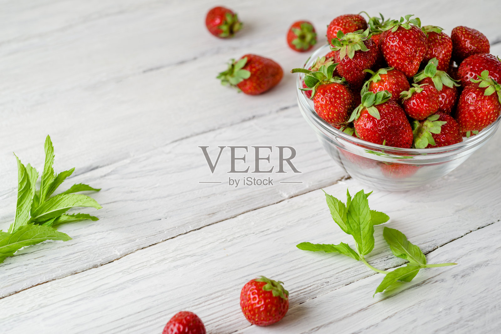 白色木桌上放着一个玻璃盘子，上面放着成熟的红草莓和绿色的薄荷叶。桌上散落着草莓。照片摄影图片