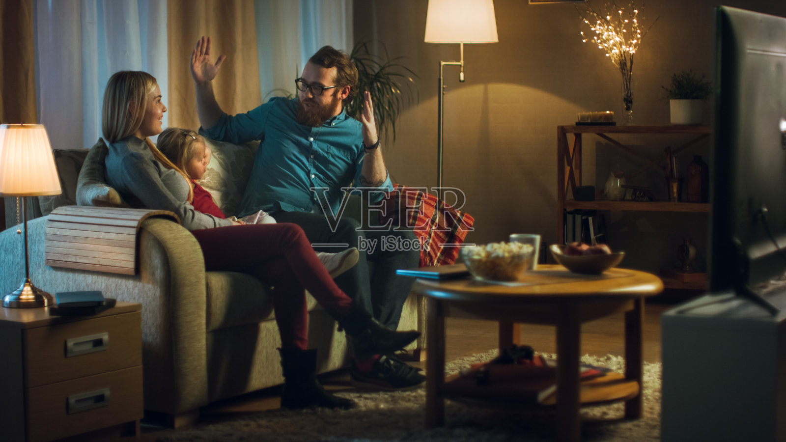 父亲，母亲和小女孩看电视的可能性不大。他们坐在舒适的客厅里的沙发上吃着爆米花。这是晚上。照片摄影图片