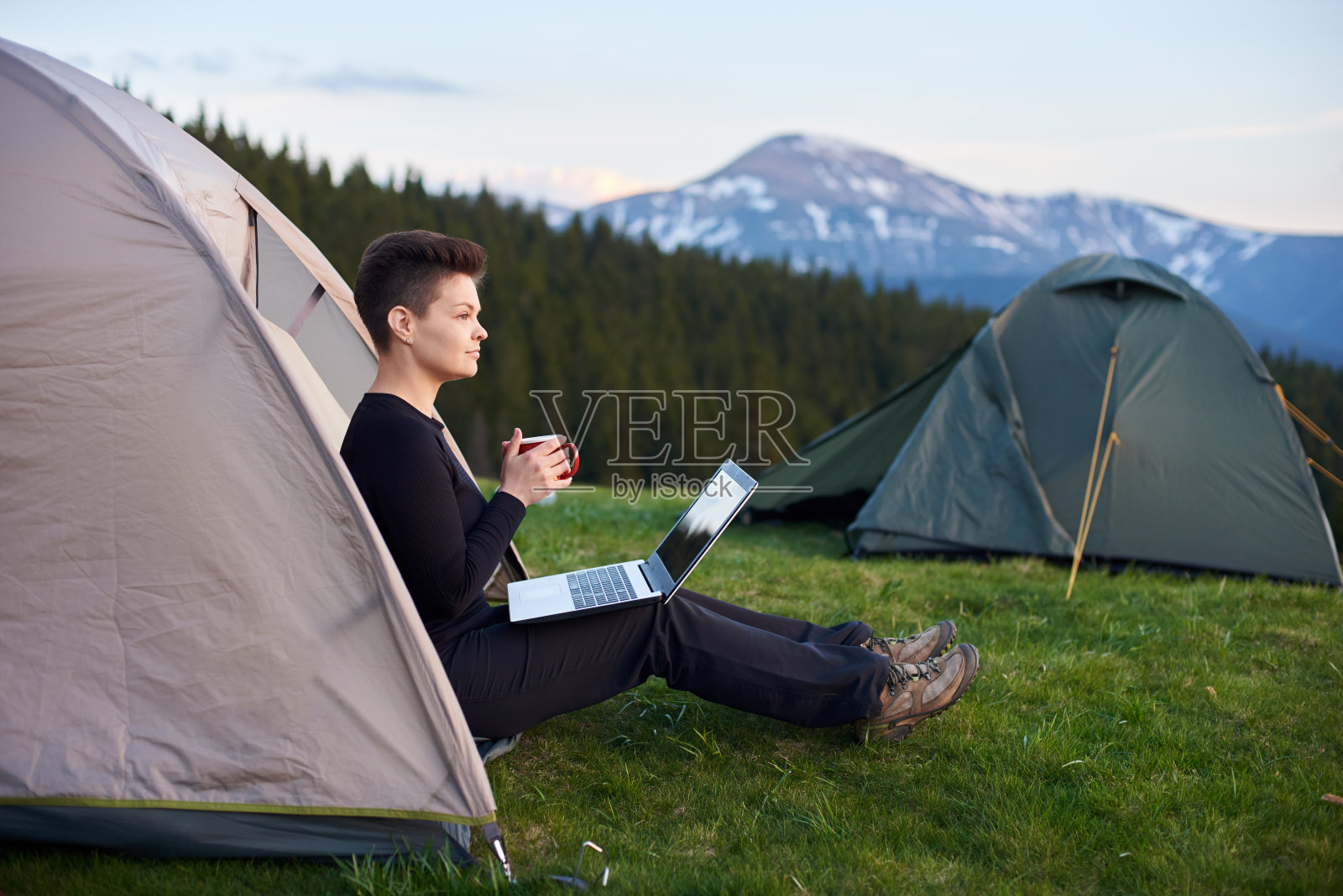 一名年轻女子在露营地的帐篷里放松，手里拿着一个杯子，在笔记本电脑上工作后，目光转向别处。背景上的山脉照片摄影图片