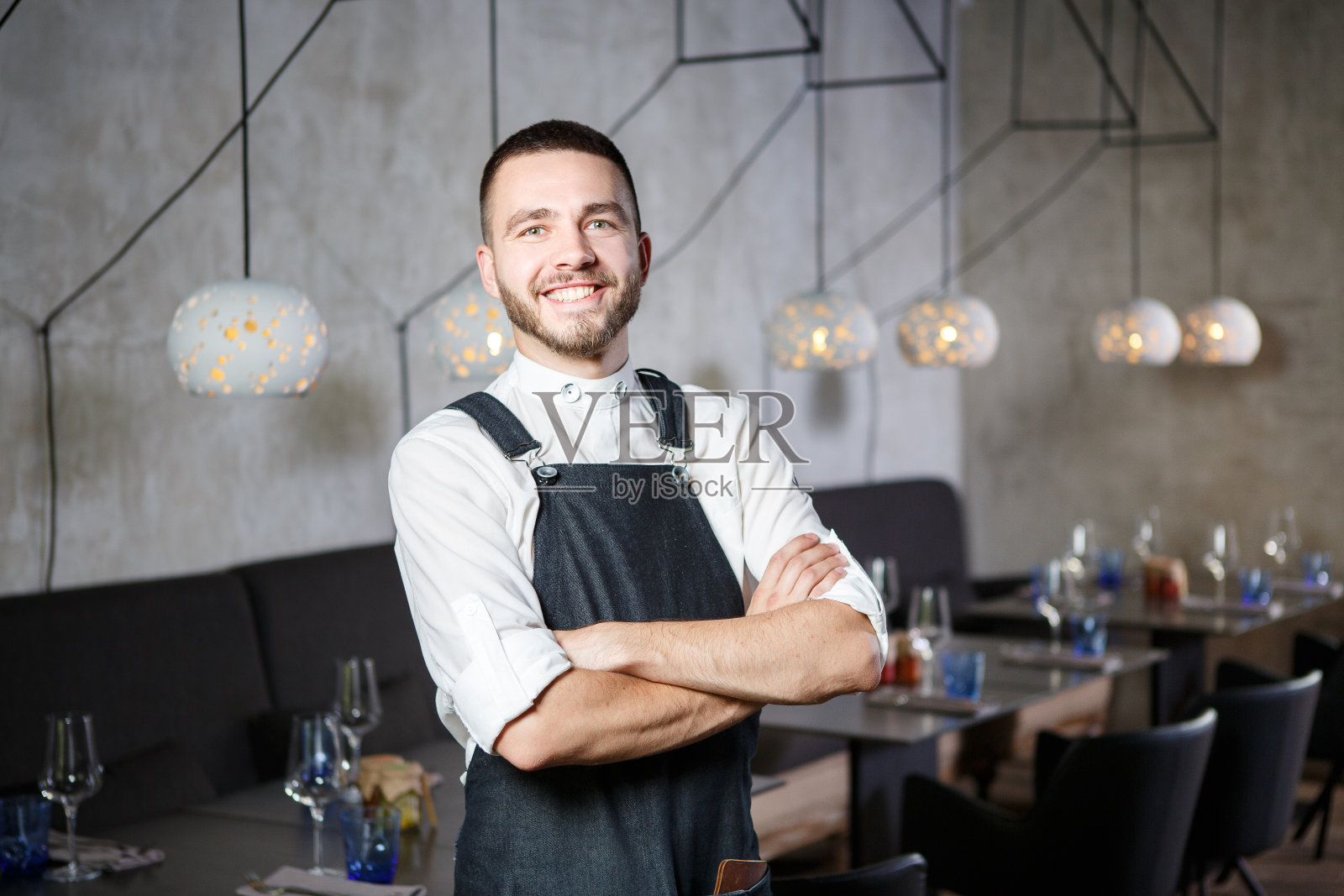 一个年轻的，微笑的服务员站在桌子旁边，拿着一杯酒。他系着围裙，看起来很自信，双臂交叉放在胸前照片摄影图片