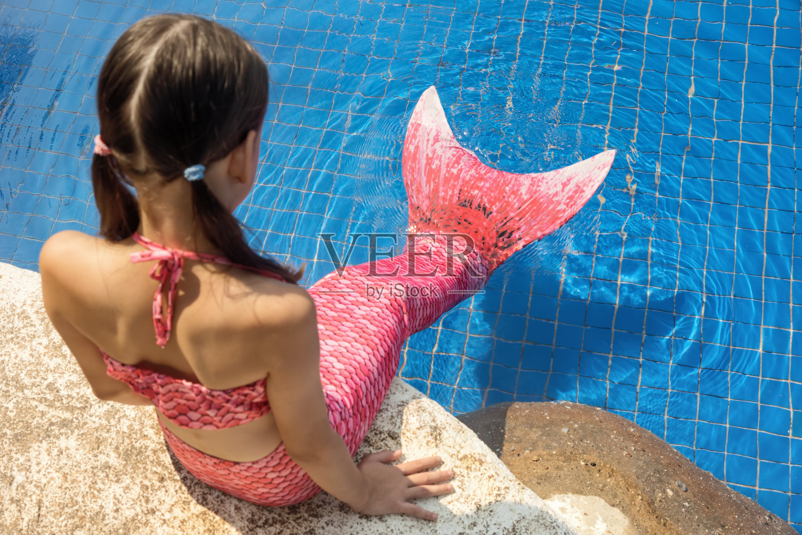 在泳池边的岩石上有一条粉红色尾巴的美人鱼女孩把脚放在水里照片摄影图片