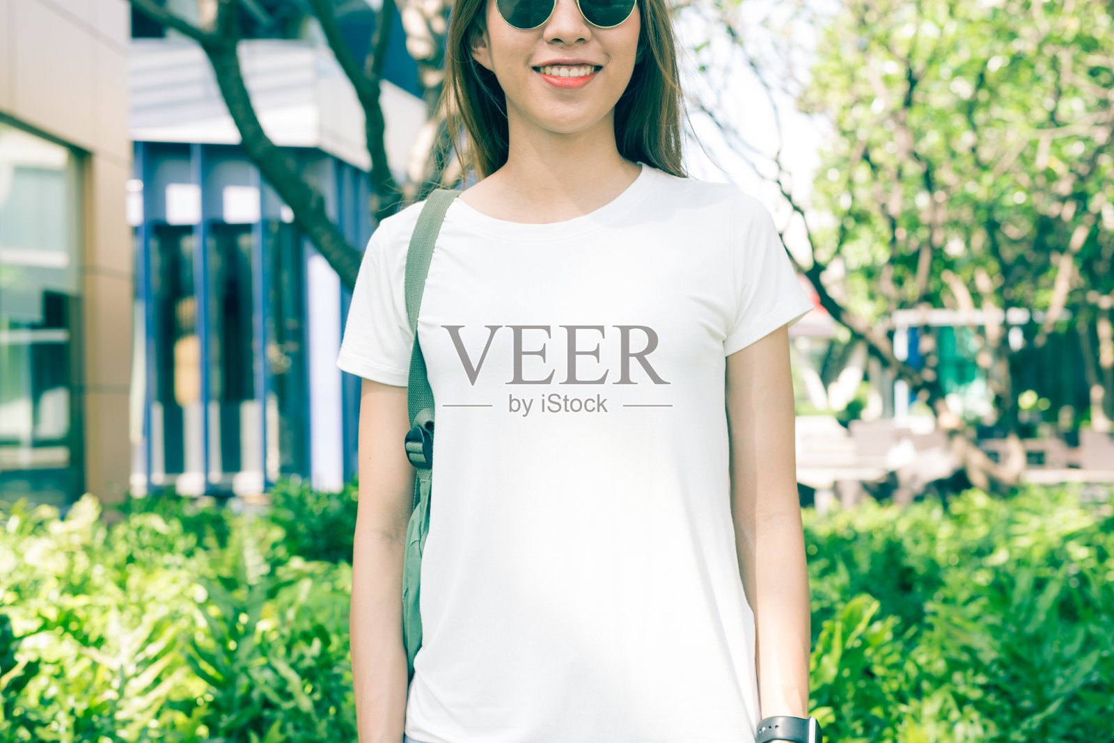 亚洲嬉皮女孩棕色长发在白色空白t恤站在街道中央。一位身着街头服饰的女性站在绿色的城市背景上。为文本或设计留出模拟空间。照片摄影图片