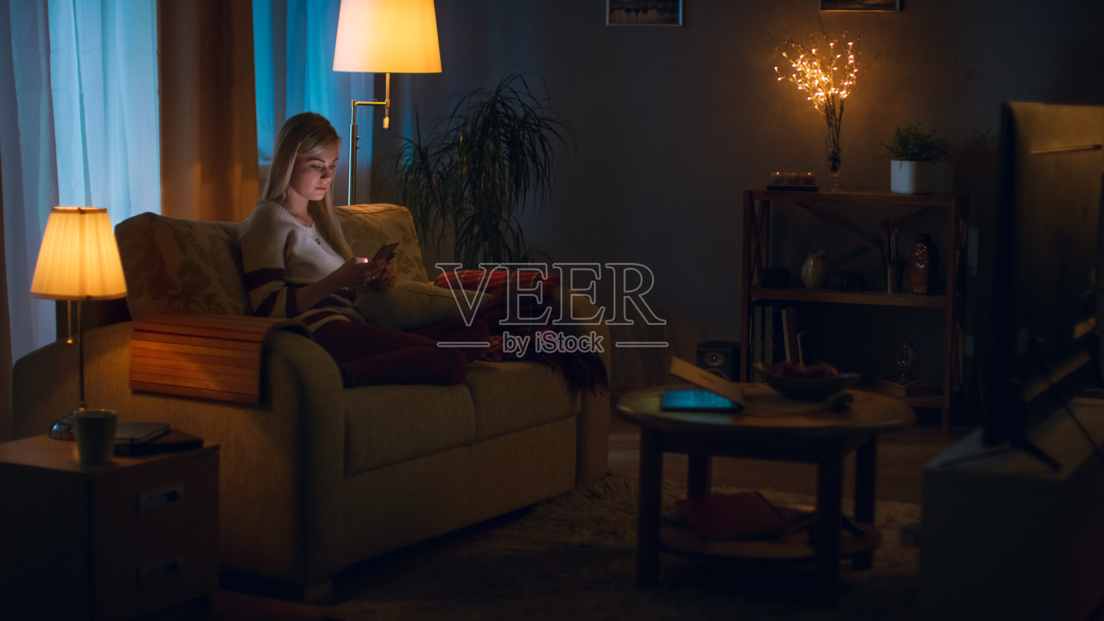 在晚上美丽的年轻女人放松在沙发上在她舒适的客厅。她使用智能手机。照片摄影图片