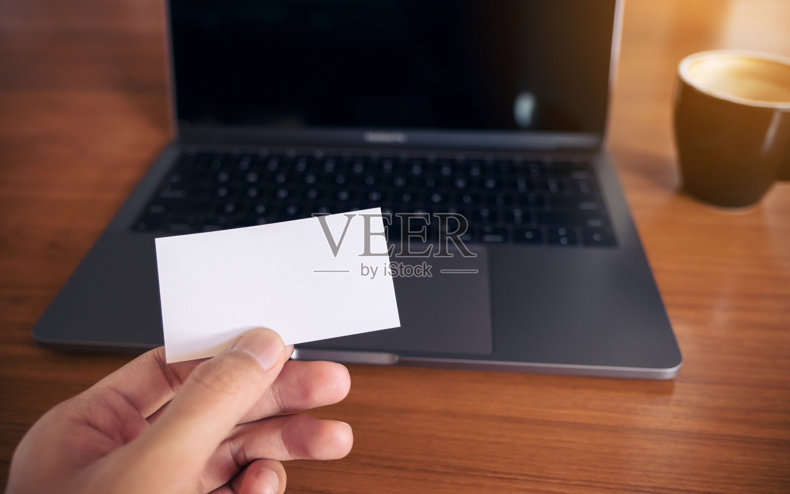 在办公室的木桌上，一只手拿着白色的空名片和笔记本电脑照片摄影图片