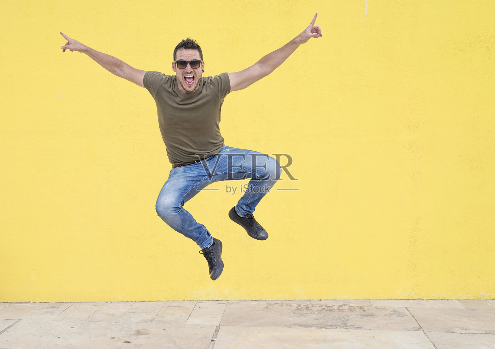 一个戴着墨镜的年轻人跳到一堵黄色的墙前。照片摄影图片