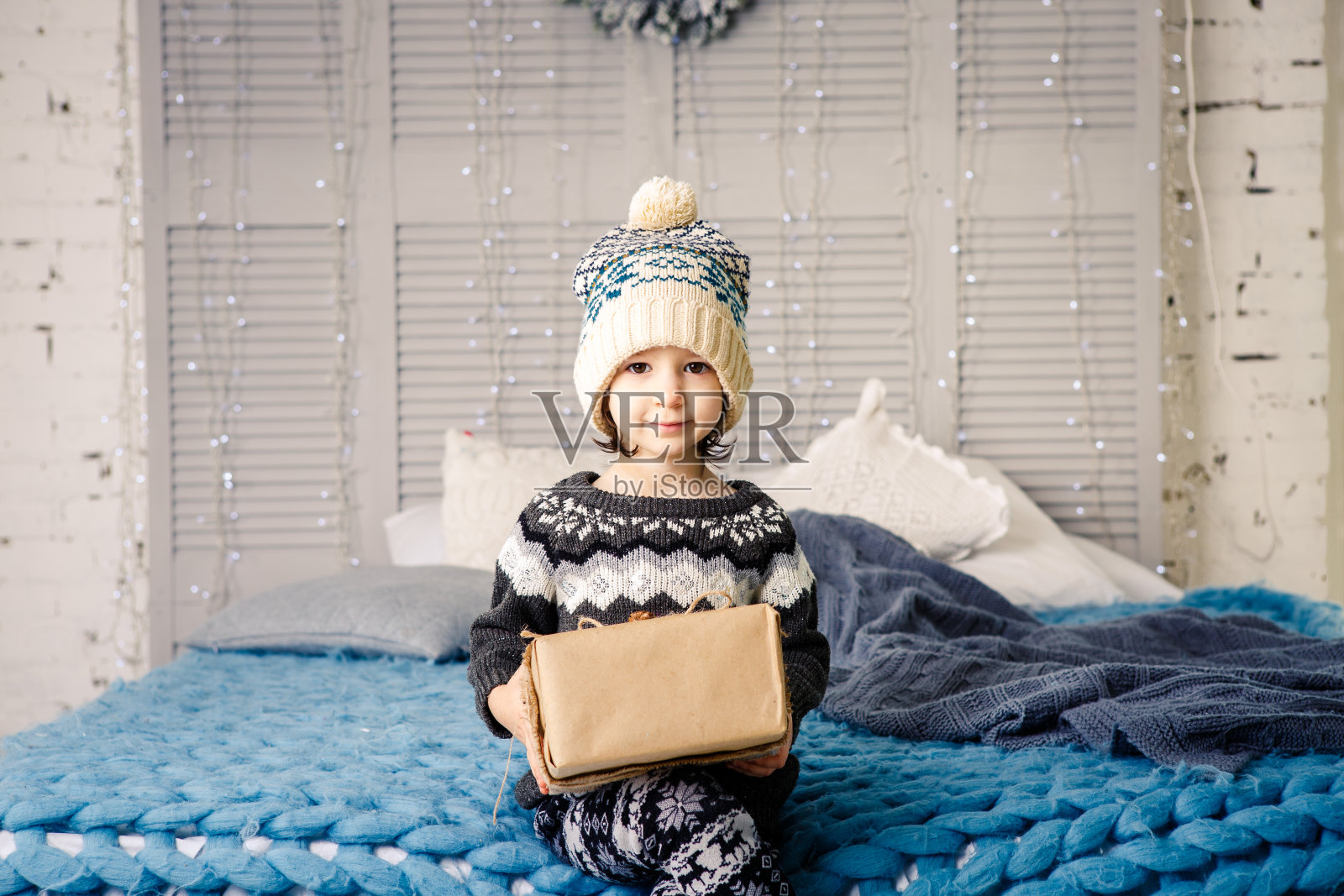 小女孩，那个穿着睡衣和帽子坐在床上的孩子，有灯泡的花环，还有用非彩色纸包着的礼品盒，蓝色针织被子上装饰着圆锥形。圣诞节的概念照片摄影图片
