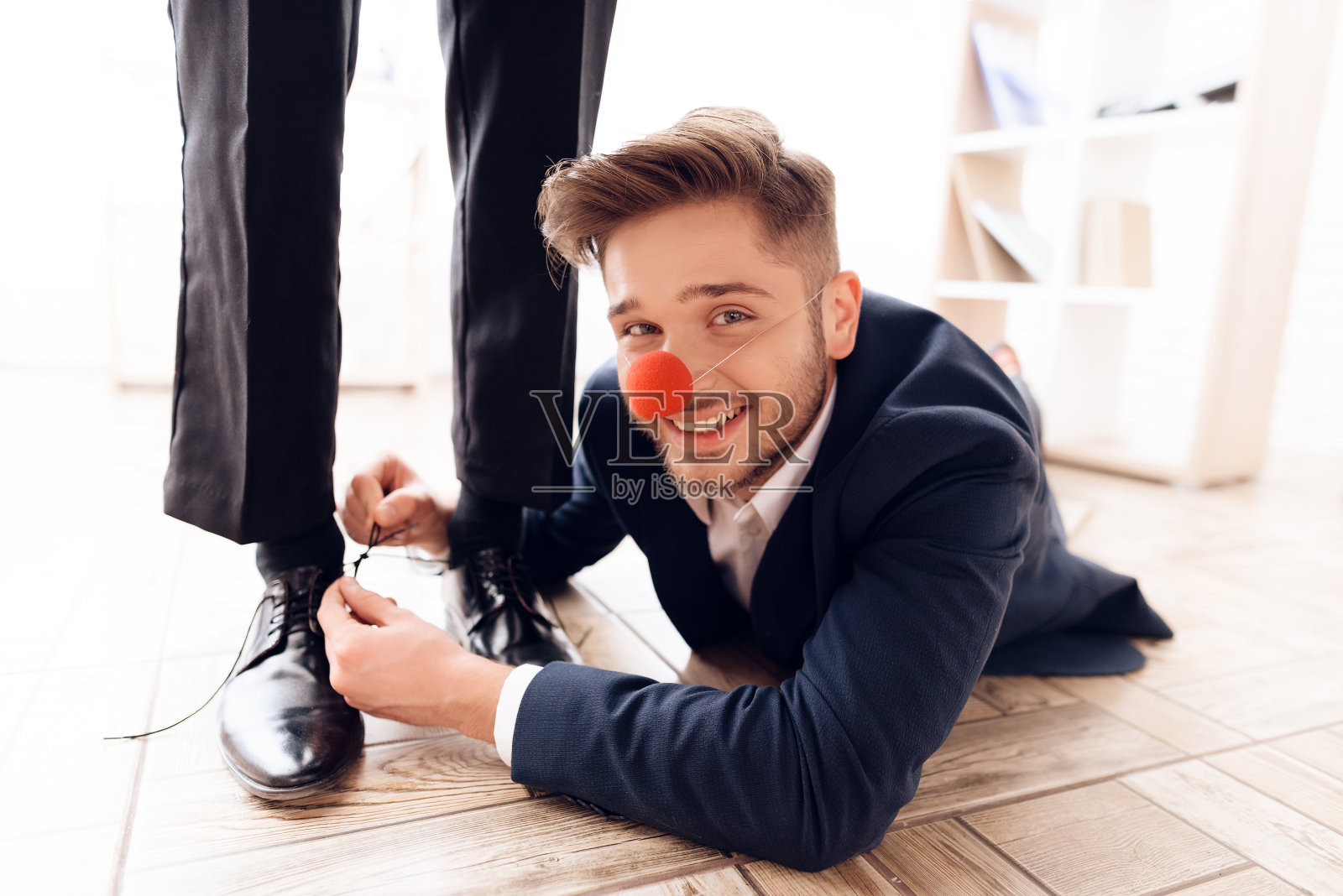 一个长着小丑鼻子的人在桌子底下给他的生意伙伴系鞋带。照片摄影图片