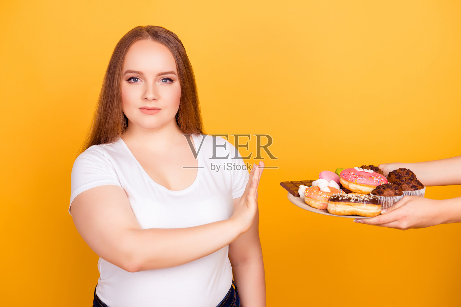 我反对吃含脂肪的食品!一个身穿白色t恤的意志坚强的女人正在拒绝吃盘子上的美味的糖果，孤立在明亮的黄色背景照片摄影图片