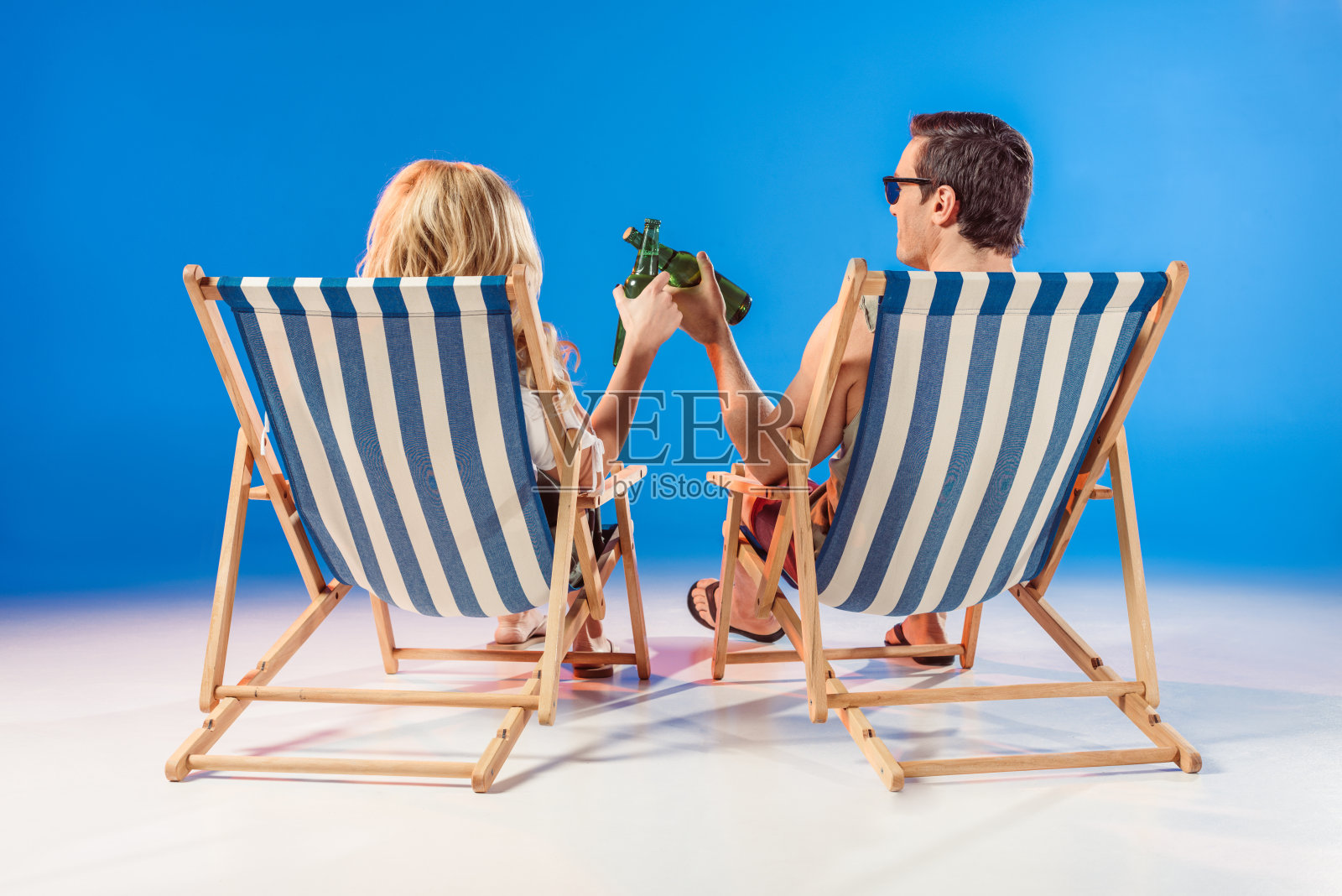 一对戴着太阳镜微笑的情侣在蓝色背景的躺椅上喝酒照片摄影图片