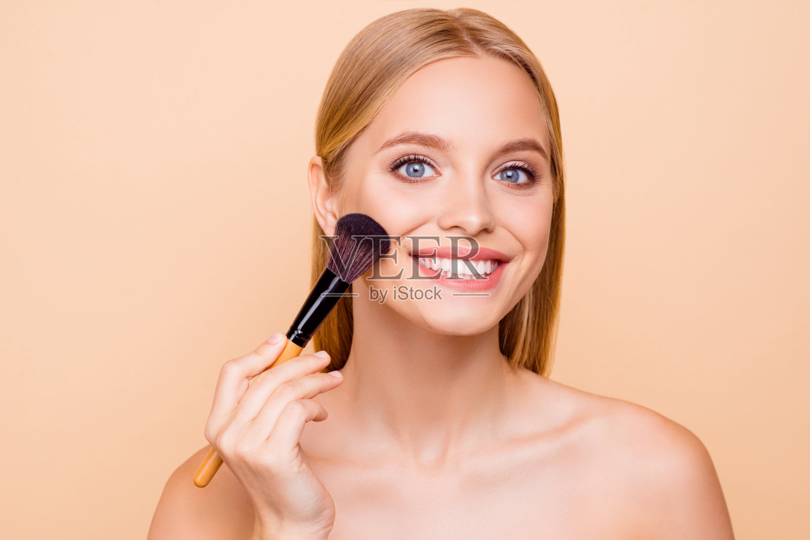迷人的漂亮开朗的正面牙齿的女人用化妆刷涂抹粉底粉在脸颊上，有完美光滑柔软的皮肤，看着相机，孤立在米色背景照片摄影图片