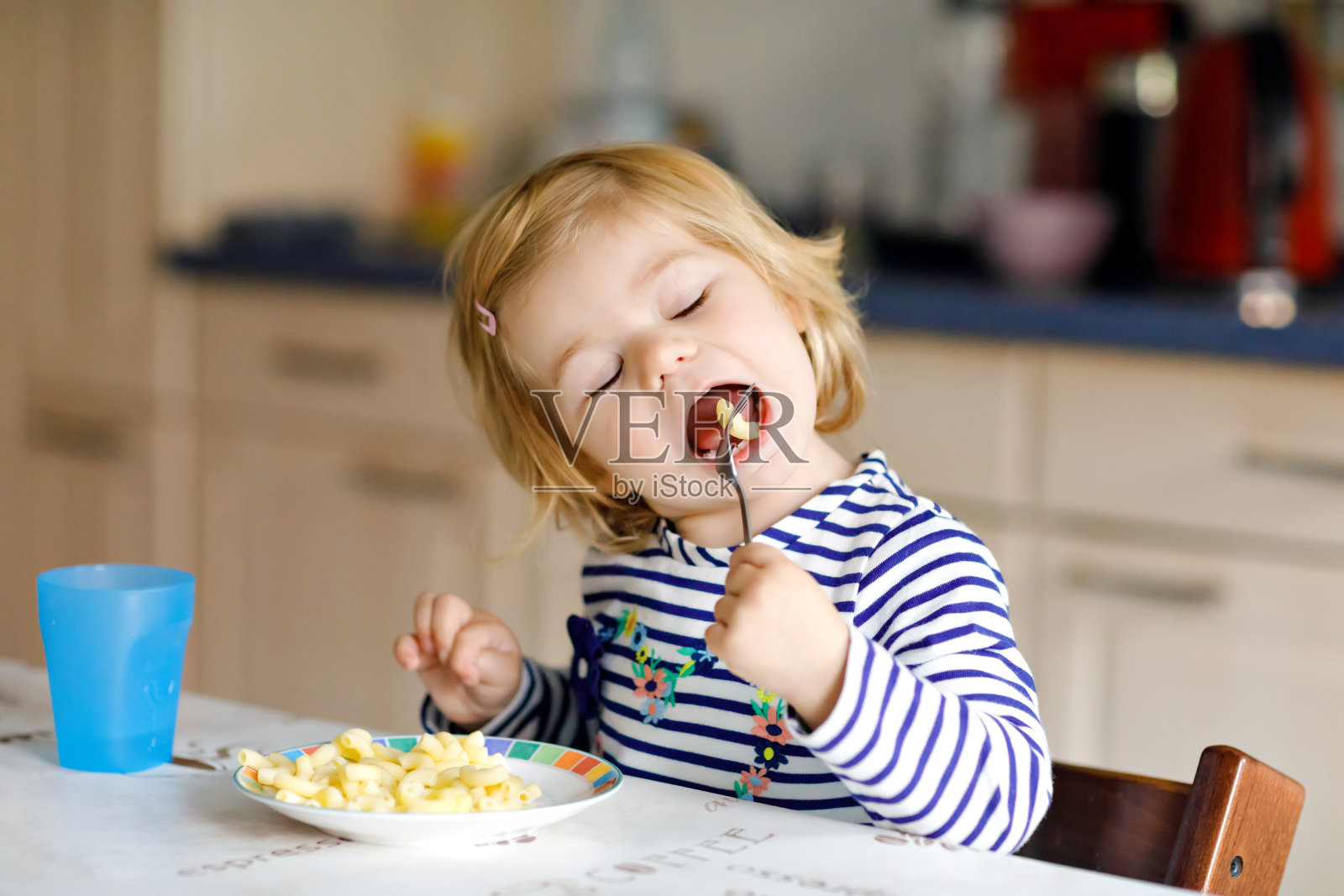 可爱的小女孩吃着勺子面条，通心粉。可爱健康的蹒跚学步的孩子，用勺子坐在高椅子上的女儿，学习在家庭厨房或托儿所自己吃饭照片摄影图片