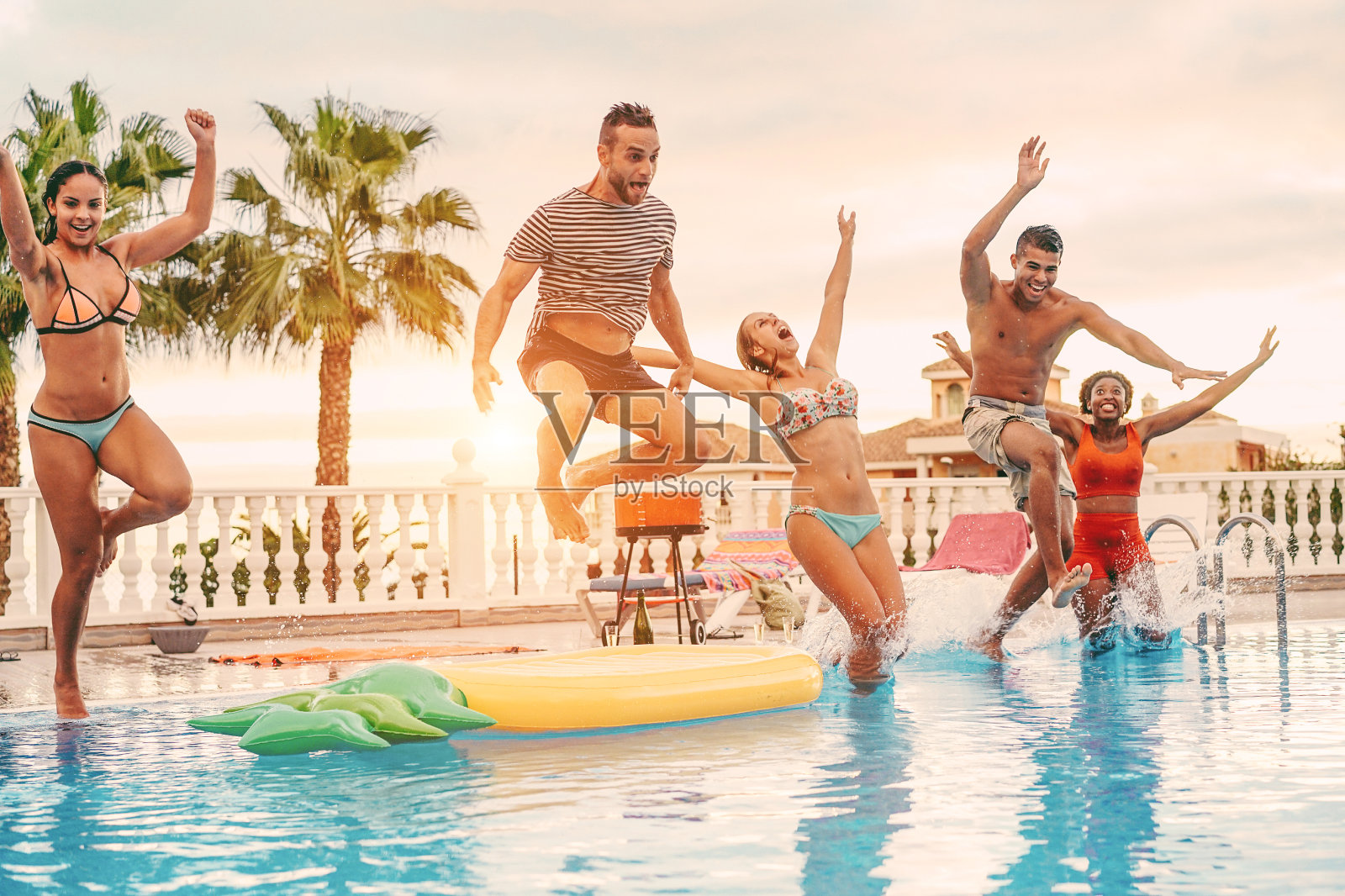 一群快乐的朋友喝在游泳池跳户外日落派对-年轻多元文化的人在热带度假的乐趣-假日，青年和友谊的概念-主要集中在左边的人照片摄影图片