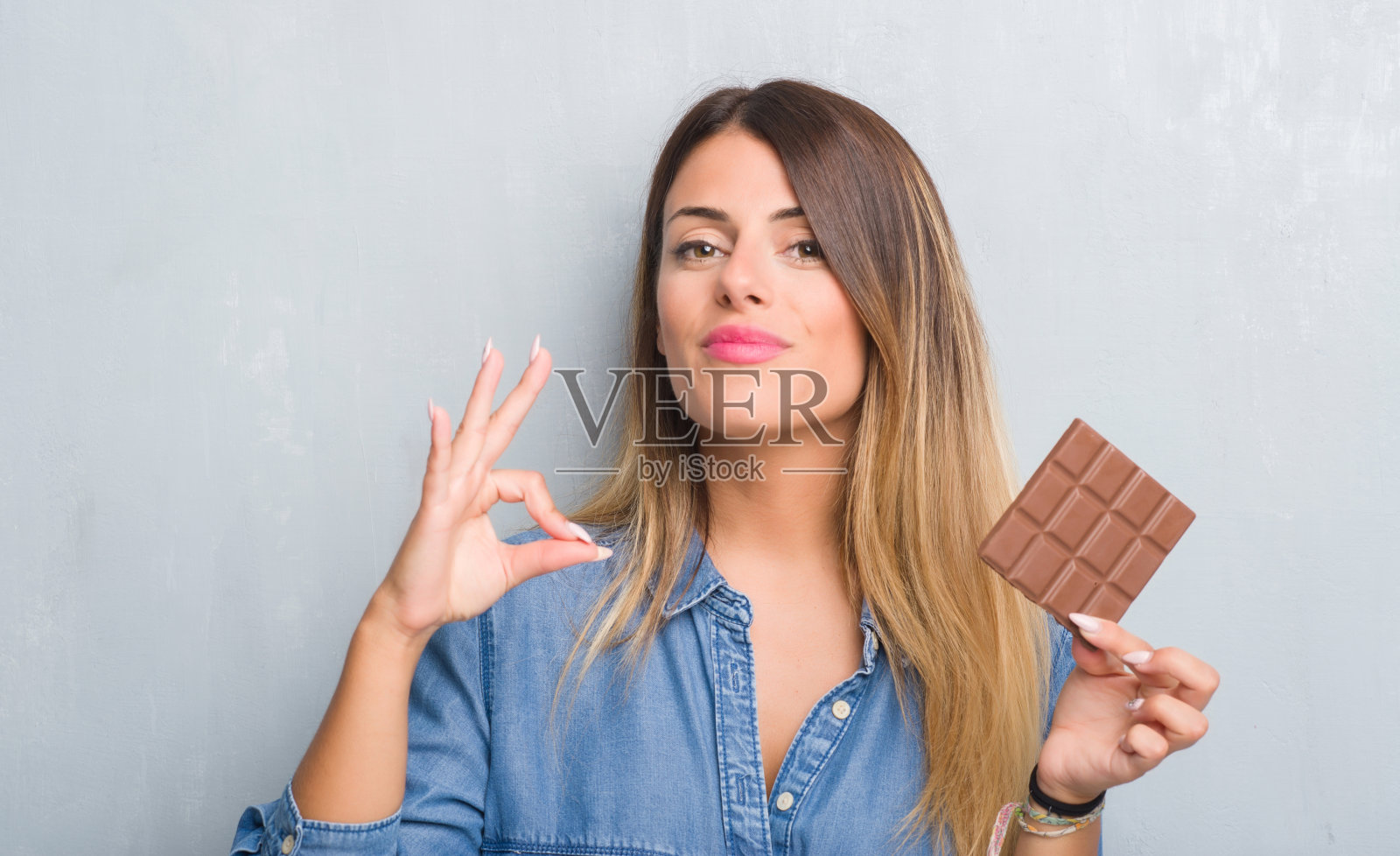 年轻的成年女子越过灰色的垃圾墙吃着巧克力棒用手指做ok的手势，很好的象征照片摄影图片