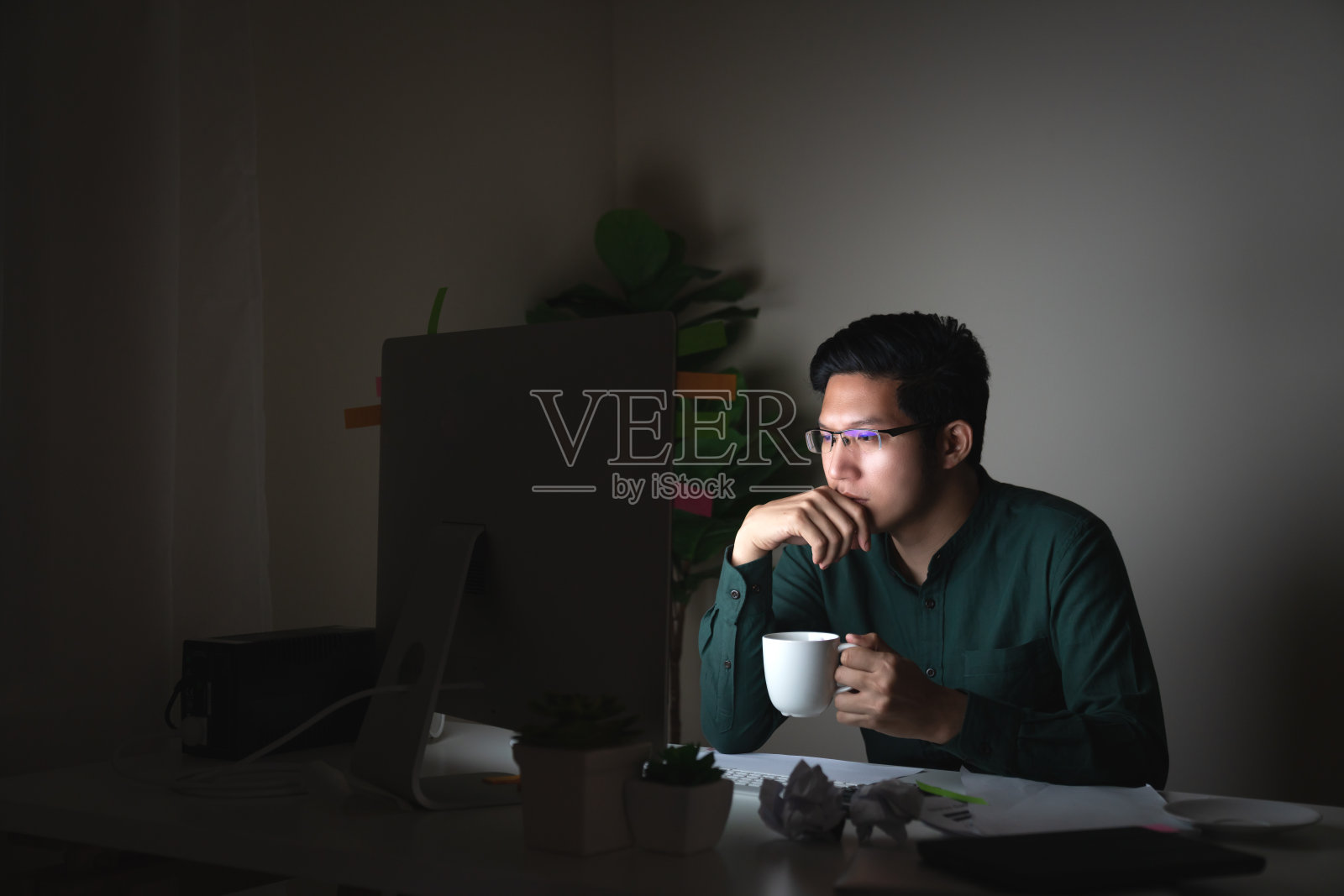 有吸引力的年轻亚洲男子喝咖啡坐在桌子上看着笔记本电脑在黑暗的深夜工作感觉认真思考和决心在家办公室努力工作的概念。照片摄影图片