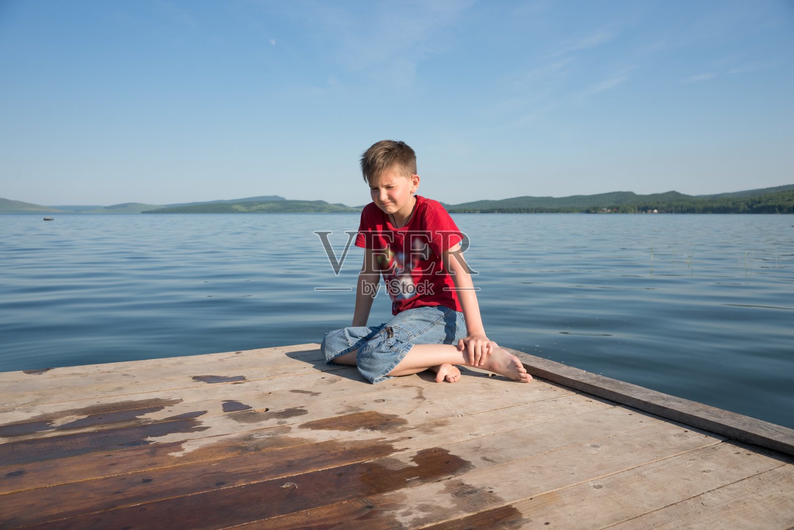 在一个阳光明媚的日子里，男孩坐在一个木码头上，对着湖面做着沮丧的鬼脸。照片摄影图片