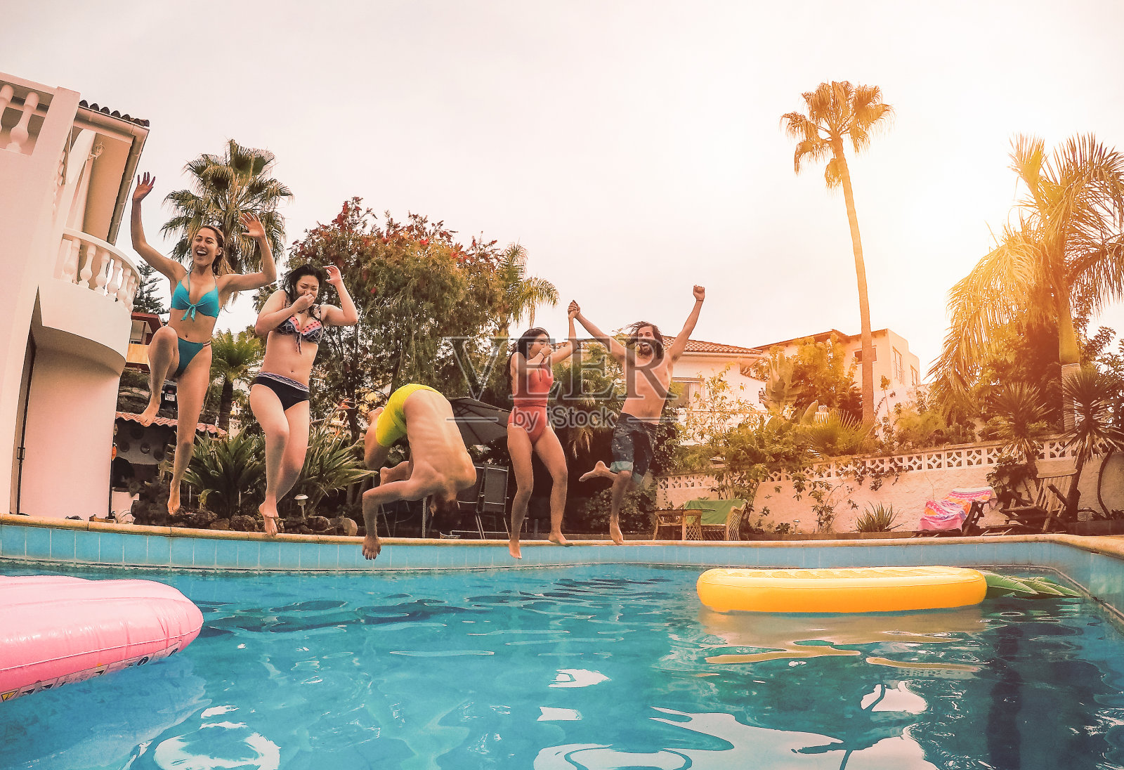 一群快乐的朋友在日落的时候跳进游泳池-千禧一代的年轻人有有趣的聚会在独家度假热带-假日，夏季，假期和青年的生活方式概念照片摄影图片