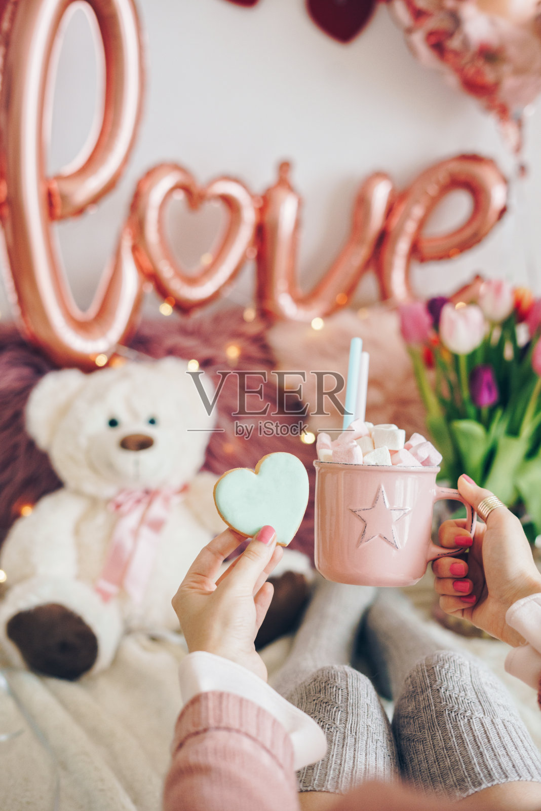 情人节概念:女人手里拿着一杯可可饮料，里面有棉花糖和一个心形饼干。气球的背景装饰。照片摄影图片