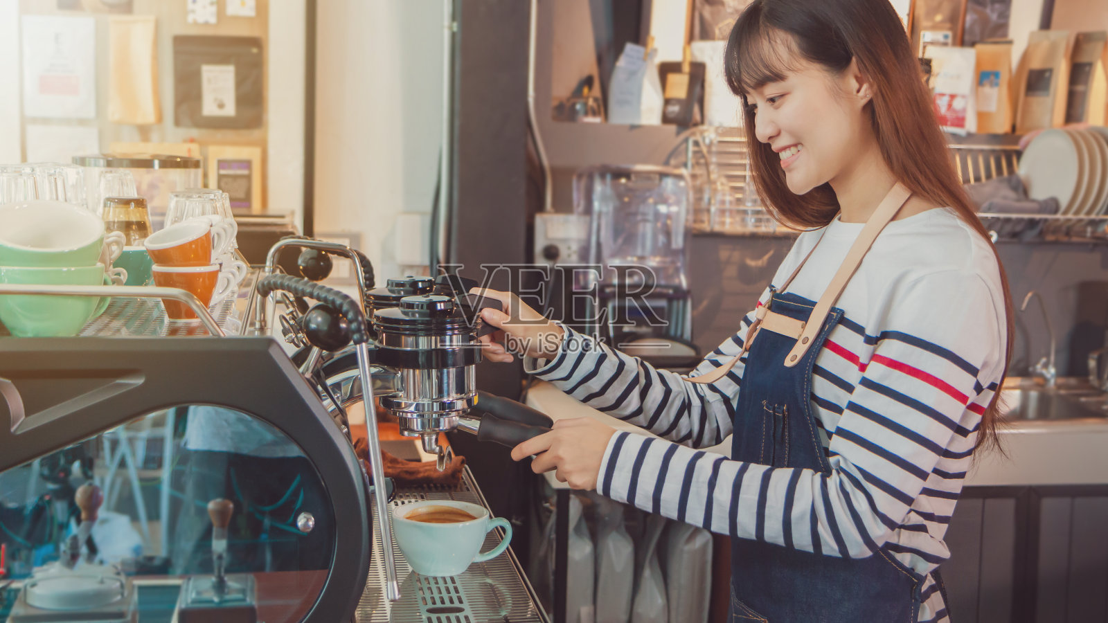 年轻美丽的亚洲女咖啡师，穿着蓝色围裙，拿着热咖啡杯，在咖啡店的吧台微笑着招待顾客。咖啡店和小型咖啡店的概念。的语气照片摄影图片