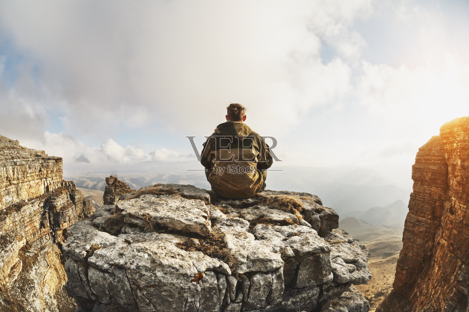 一个年轻人坐在一块被悬崖环绕的岩石上，位于云层之上的一个很好的位置，背景是云、山谷、田野和山脉照片摄影图片