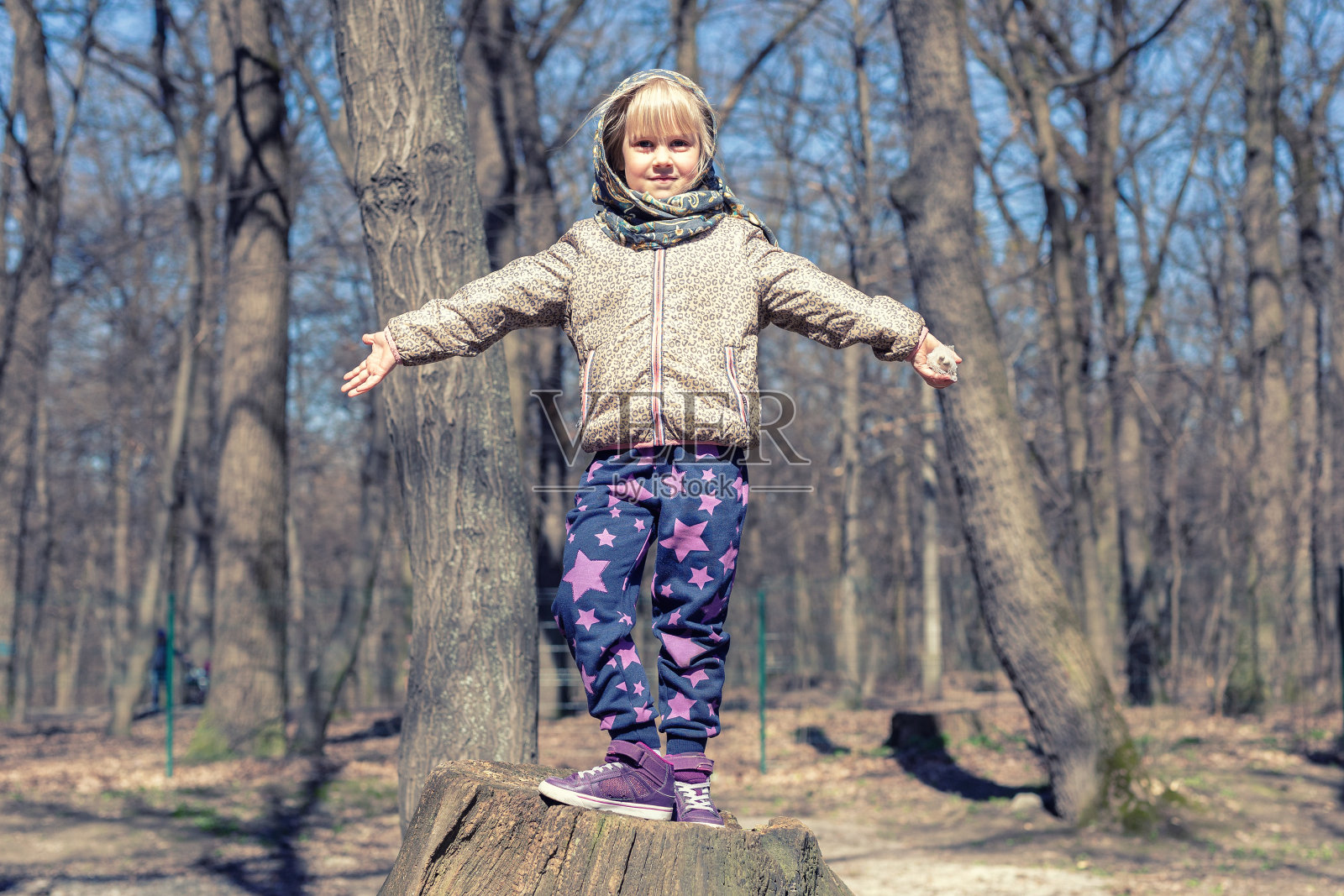 可爱的金发小女孩在户外玩。穿着休闲运动服和头巾的孩子在森林里从树桩上跳得很高照片摄影图片