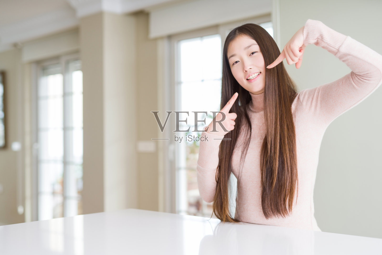 美丽的亚洲女人穿着休闲毛衣在白色的桌子上微笑自信的显示和指着手指，牙齿和嘴。健康的概念。照片摄影图片