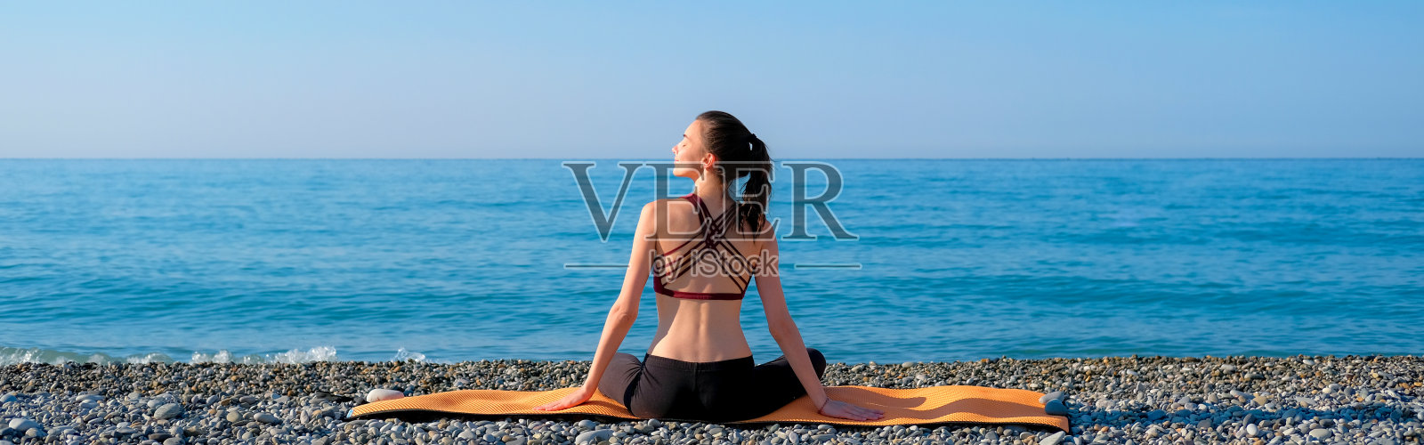 年轻苗条的女人穿着紧身运动服坐在橙色的瑜伽垫上，在海边的卵石滩户外练习。自然瑜伽概念照片摄影图片