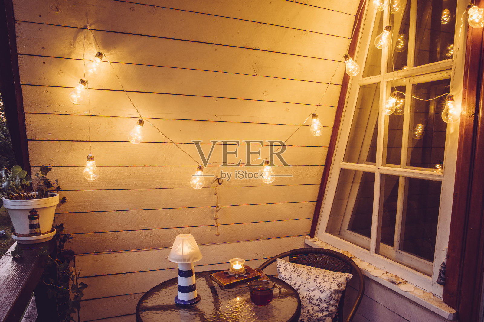 可爱的复古木制航海风格阳台视图与小花园桌椅和装饰串派对灯泡在晚上打开。照片摄影图片