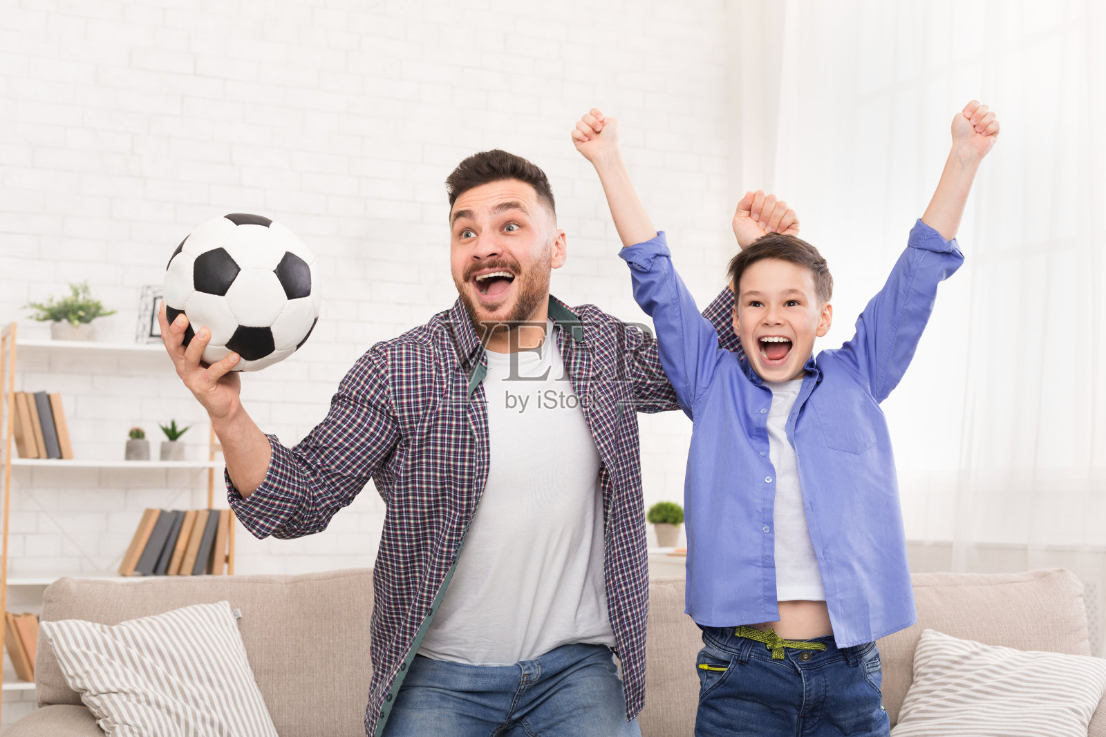 激动的爸爸和儿子拿着足球欢呼，在家里庆祝进球照片摄影图片