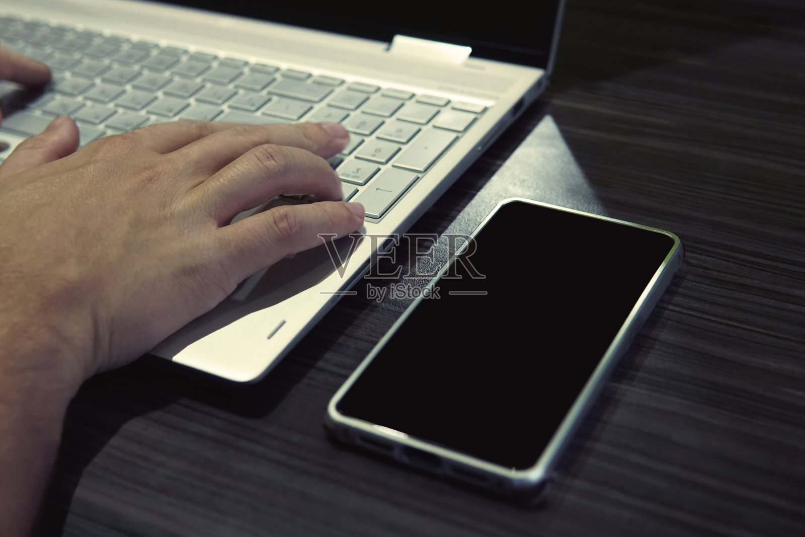 一名男子坐在笔记本电脑和智能手机后面，手握键盘。黑屏手机放在桌子上，靠近笔记本，男性的手放在键盘上。深夜工作的自由职业者。网上业务照片摄影图片