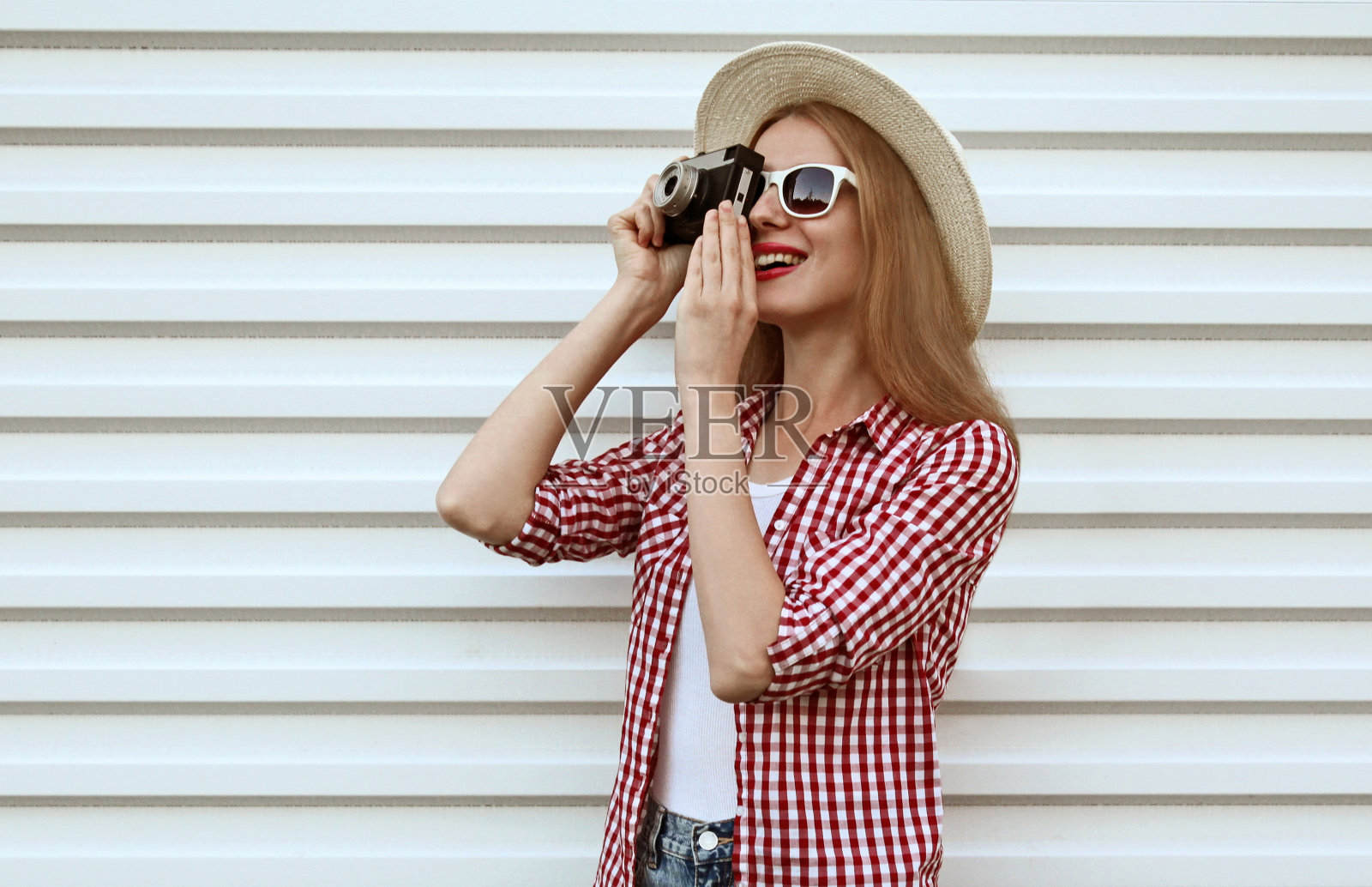 微笑的年轻女性摄影师与老式的电影相机戴着夏季圆形草帽，白墙背景上的格子衬衫照片摄影图片