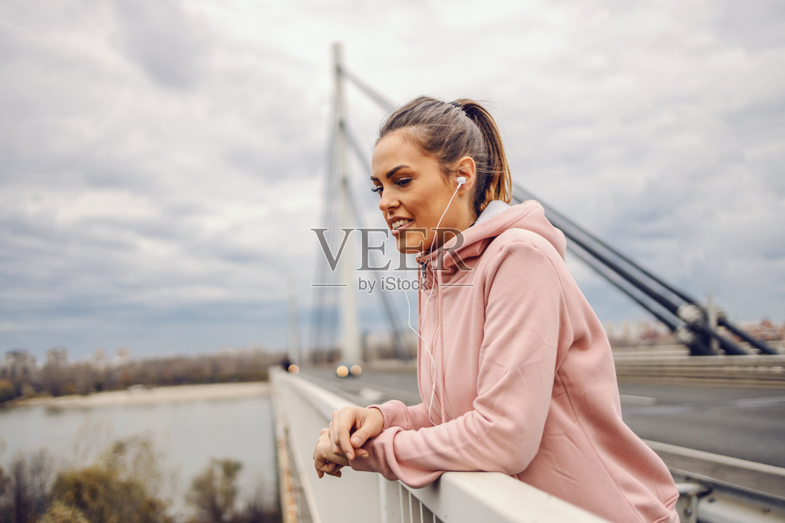 美丽的年轻女运动员倚在桥栏杆上休息，听着音乐，看着河流。城市生活的概念。照片摄影图片