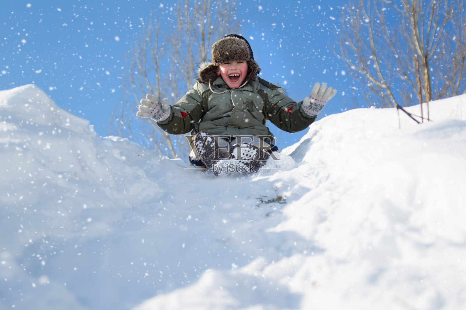 冬天的一天，一个小男孩戴着一顶带头巾的帽子走在白雪覆盖的山上。用雪橇滑雪照片摄影图片