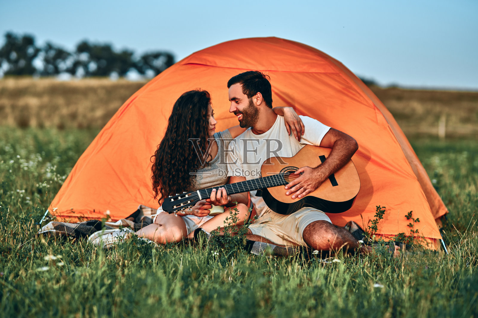 一个微笑的年轻男子为他的女朋友坐在露营帐篷弹吉他的肖像照片摄影图片