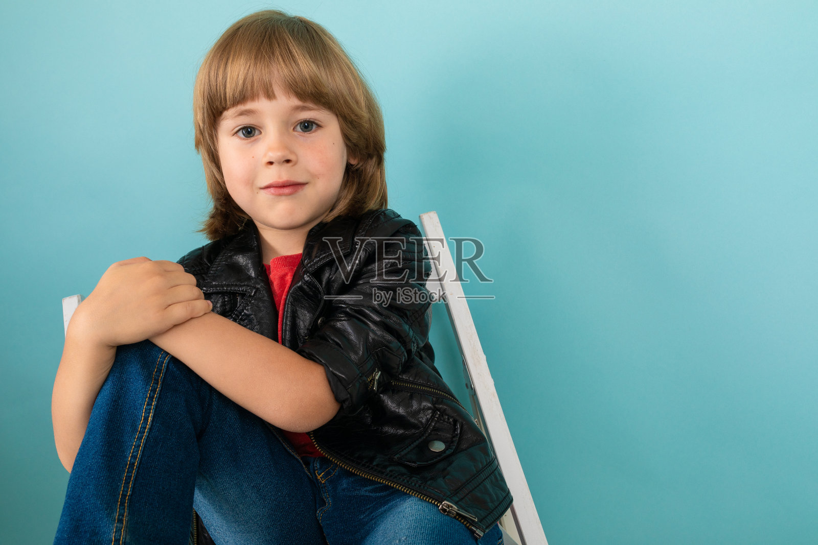 一个十几岁的白人男孩坐在一张白色的椅子上，抱着一个膝盖，照片在蓝色的背景上显得孤立照片摄影图片