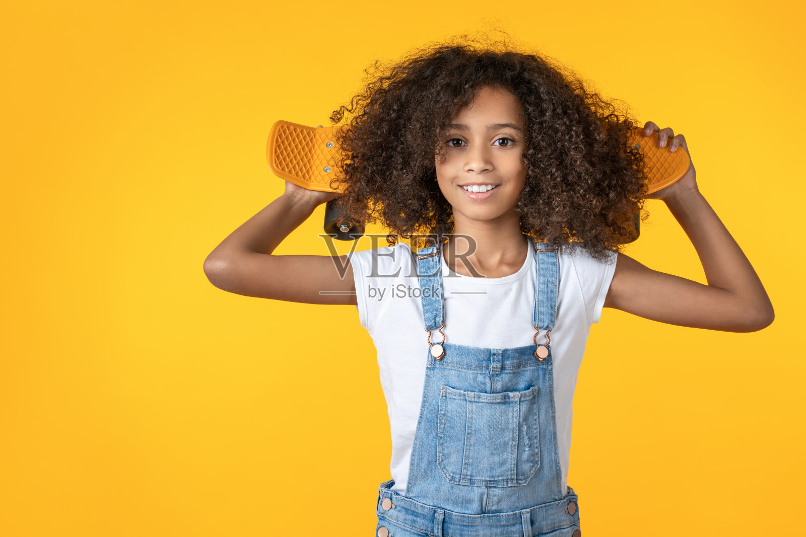 快乐的十多岁的女孩拿着滑板在她的肩膀上孤立在黄色的背景照片摄影图片