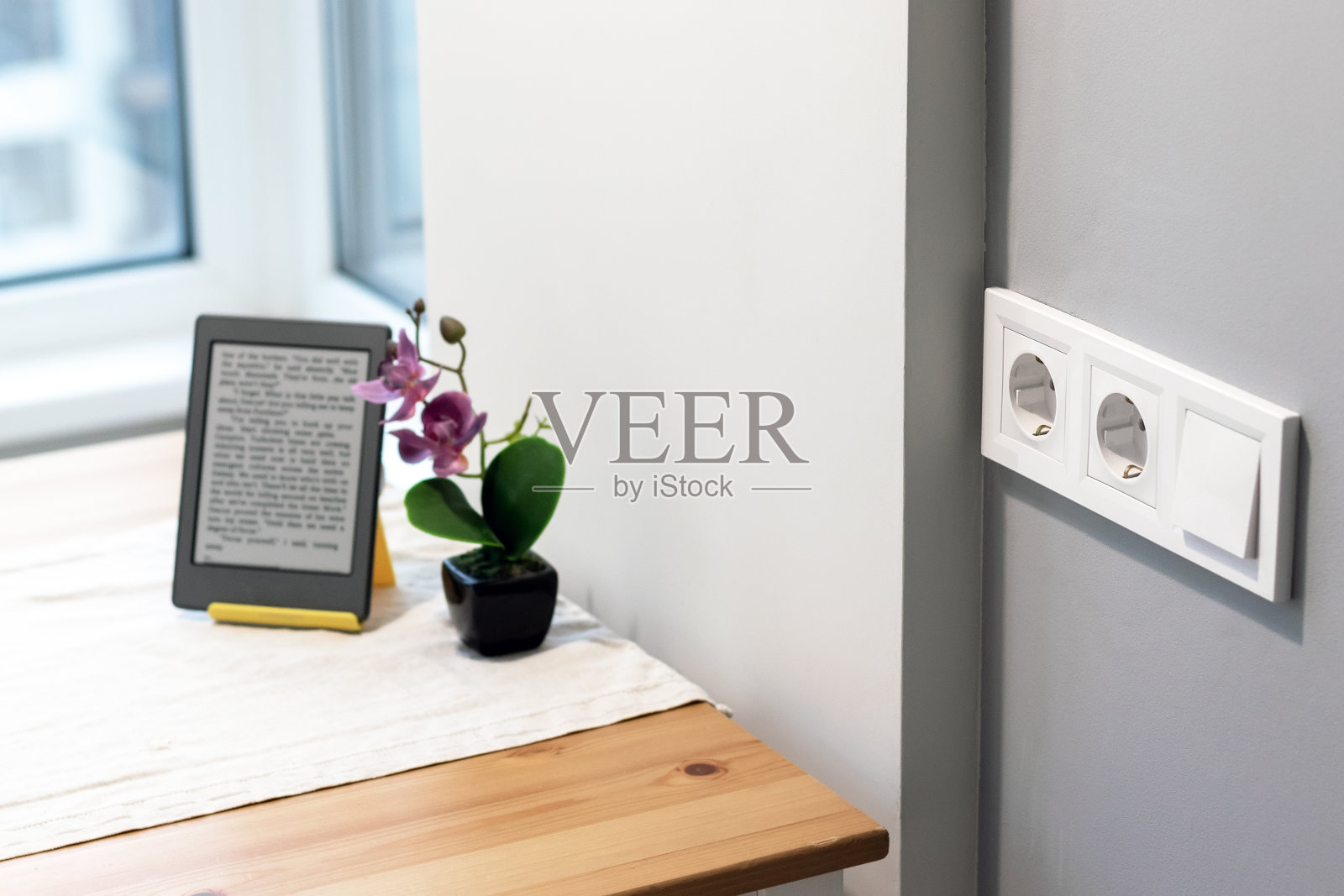 一组白色的欧洲电源插座和一个开关位于一间浅色现代厨房的灰色墙壁上，窗边的桌子上放着鲜花和电子书。有选择性的重点照片摄影图片