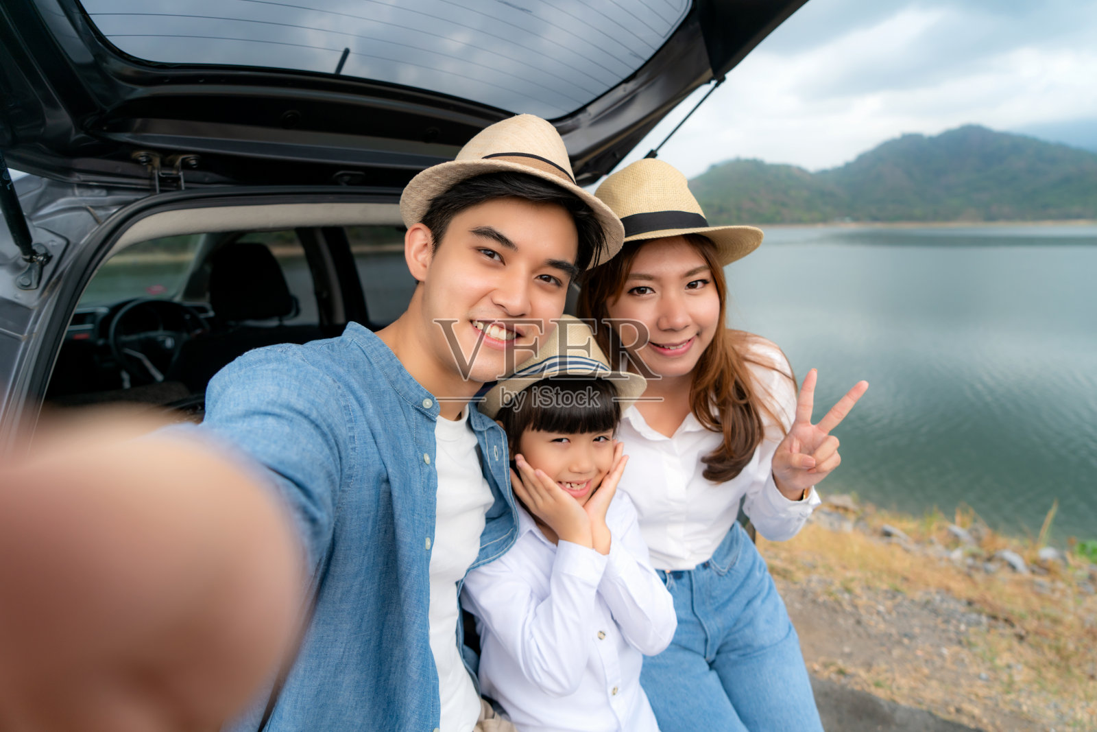 亚洲家庭坐在汽车与父亲，母亲和女儿自拍与湖泊和山景通过智能手机在假期一起度假。幸福的家庭时间。照片摄影图片