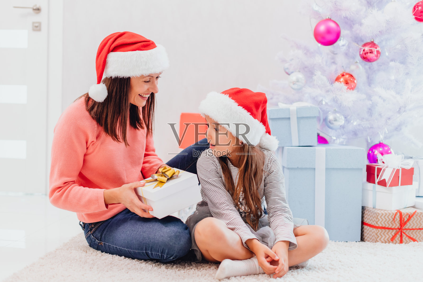 妈妈戴着圣诞老人的帽子要给她可爱的女儿送一份礼物，女儿正惊讶地看着盒子，用月开了。照片摄影图片
