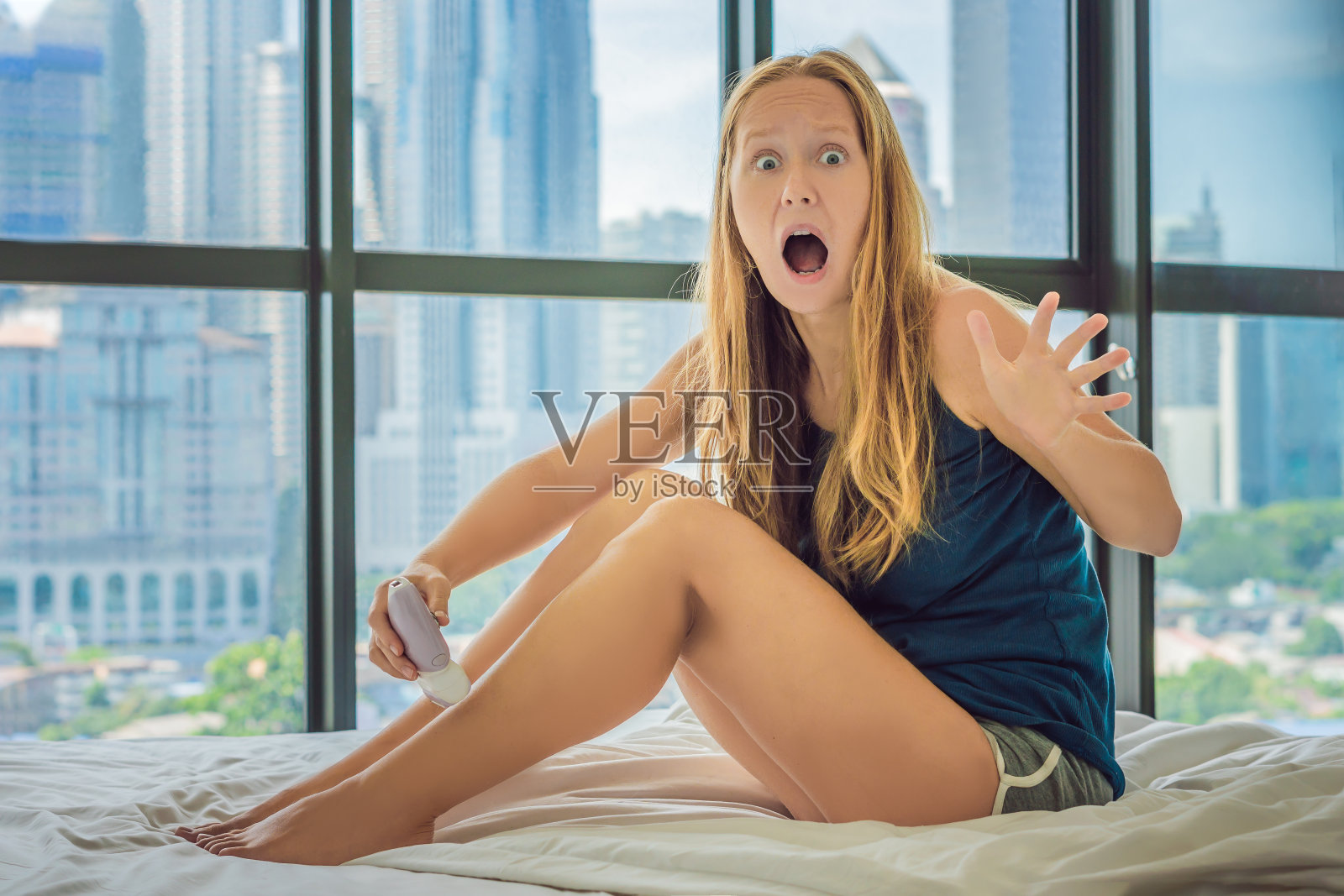 年轻女子坐在床上，在做脱毛与脱毛腿，是痛苦的。在一扇俯瞰大城市的窗户的背景上，摩天大楼照片摄影图片